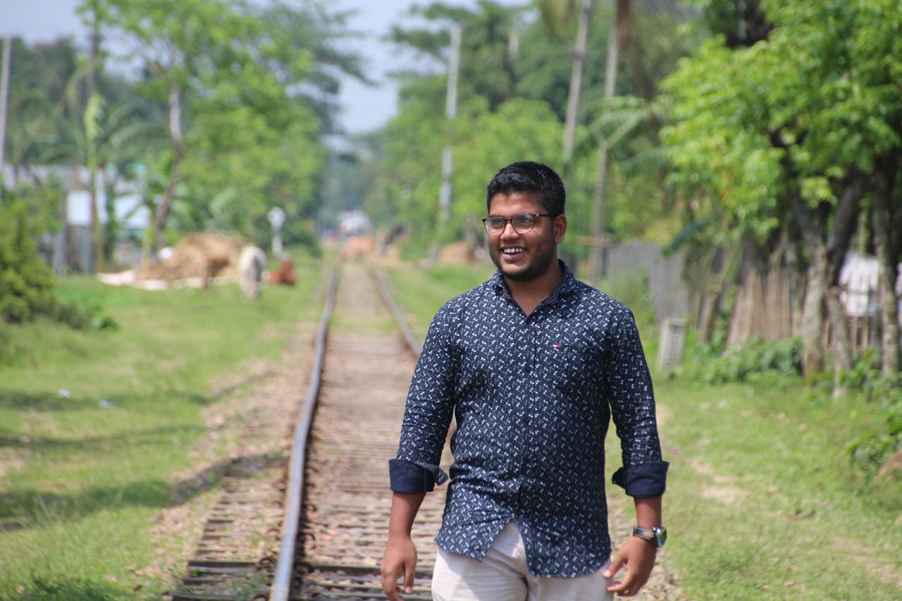 Smiling man casually walking on railway tracks surrounded by lush greenery in Jamalpur, Bangladesh.